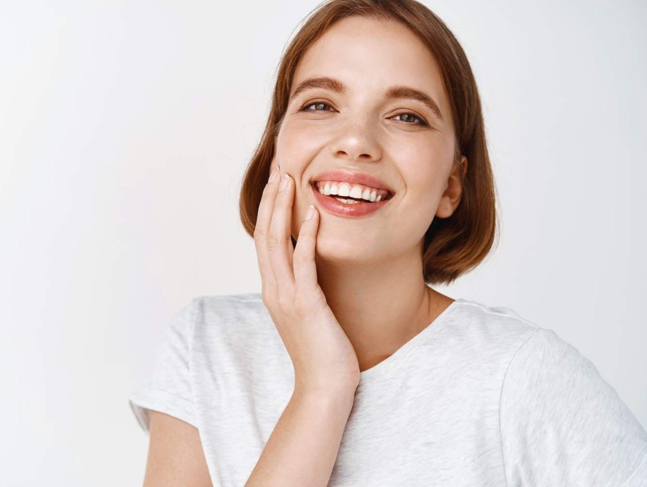 belleza-primer-plano-mujer-feliz-pelo-corto-tocando-piel-facial-limpia-fresca-sonriendo-mostrando-dientes-cara-perfectos-pie-contra-pared-blanca-